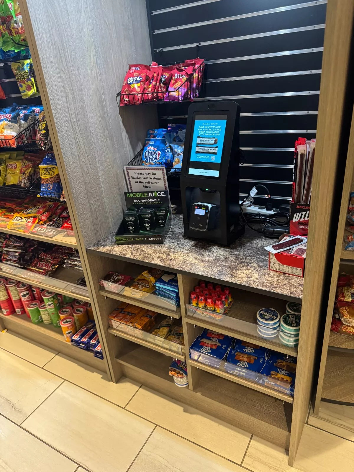 Self-service kiosk in a convenience store with snacks and drinks on shelves.